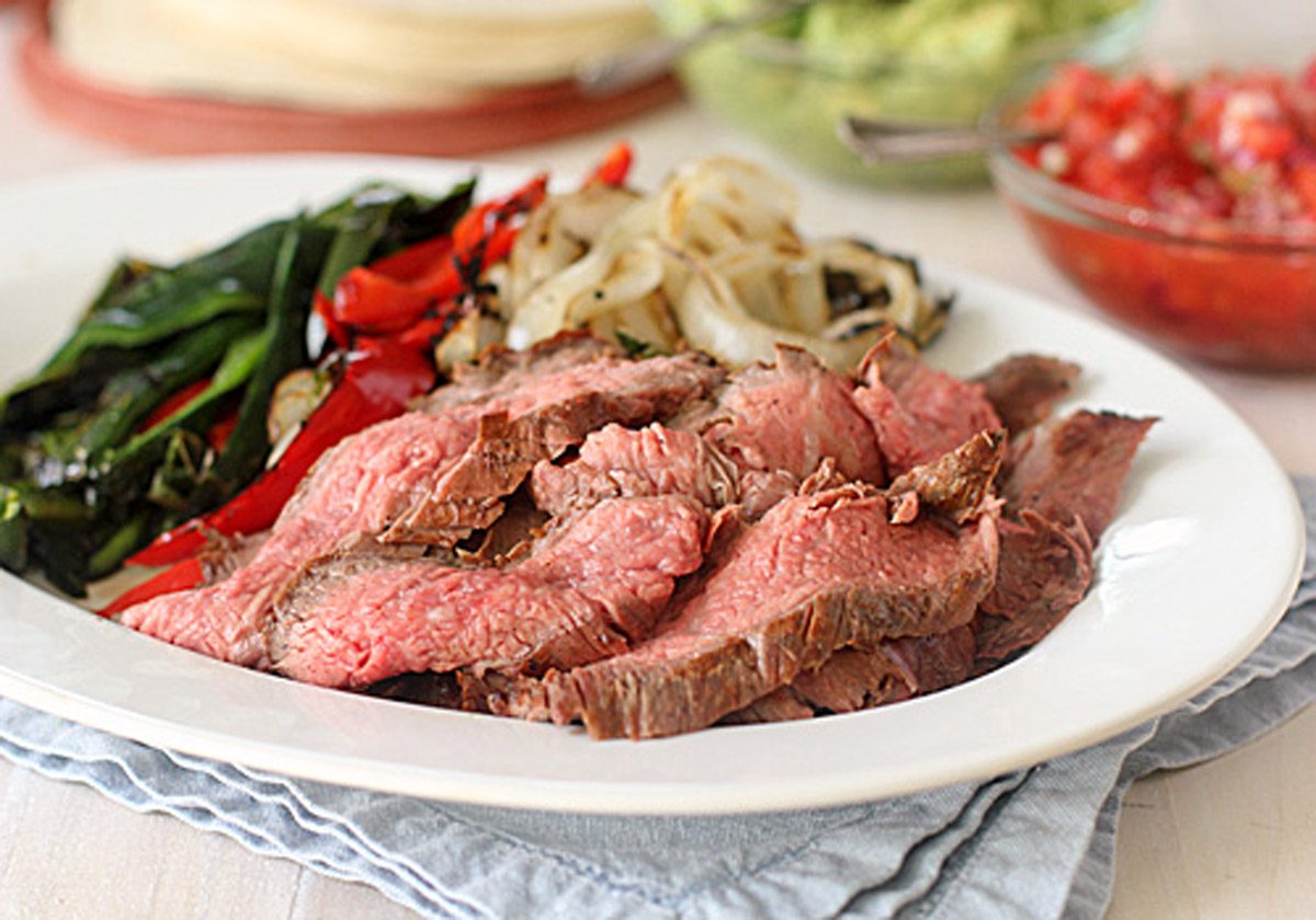 A plate of sliced grilled steak served with sautéed onions, red bell peppers, and green vegetables, sitting on a folded blue napkin, with guacamole and salsa in the background.