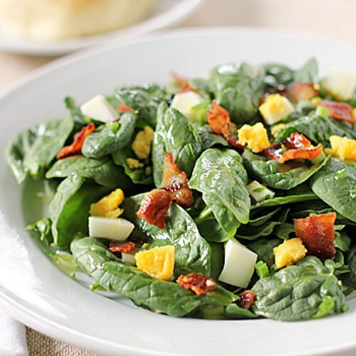A white plate of fresh spinach salad topped with chopped hard-boiled eggs and crispy bacon pieces, placed on a light tablecloth with a fork and knife beside it.
