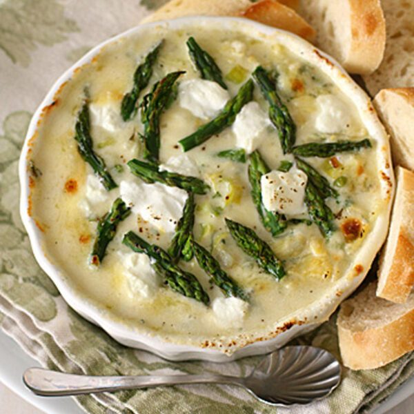 A small white dish filled with a baked creamy dip topped with asparagus tips and dollops of cheese, served on a floral napkin with slices of baguette and a small spoon on the side.
