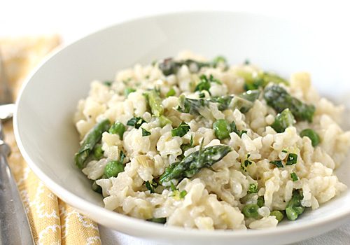 A white bowl filled with creamy risotto mixed with green asparagus pieces and peas, garnished with fresh herbs, sits on a patterned yellow napkin.