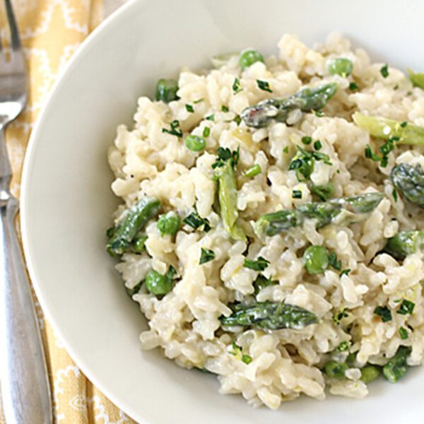 A white bowl filled with creamy risotto, green asparagus pieces, and peas, garnished with chopped herbs. A fork and knife rest on a yellow patterned napkin beside the bowl.
