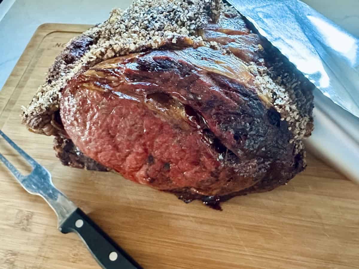 A cooked Standing Rib Roast with Au Jus, boasting a crispy, seasoned crust, rests on a wooden cutting board beside a carving fork and a large knife.