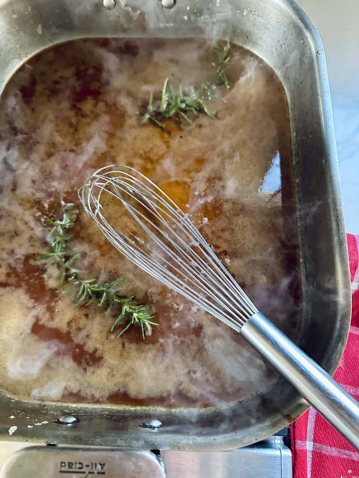 A metal whisk rests in a pan of simmering brown gravy for Standing Rib Roast with Au Jus, with sprigs of rosemary floating on top; a red and white towel is nearby.