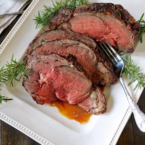 A white platter with slices of medium-rare Standing Rib Roast with Au Jus, garnished with sprigs of fresh rosemary. A serving fork rests beside the meat, and a small dish of sauce is in the background.