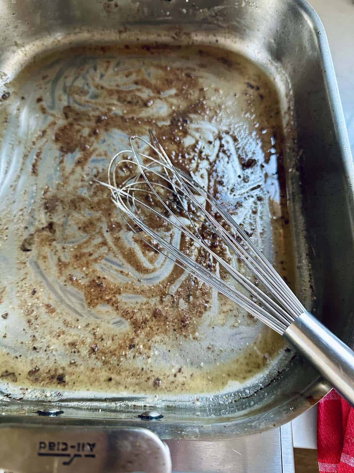 A metal whisk rests in a stainless steel pan with browned bits and melted butter, hinting at the beginnings of au jus for a Standing Rib Roast.