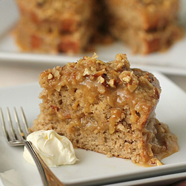 A slice of moist cake topped with chopped nuts and caramel glaze sits on a white plate with a dollop of cream and a fork; the rest of the cake is in the background.