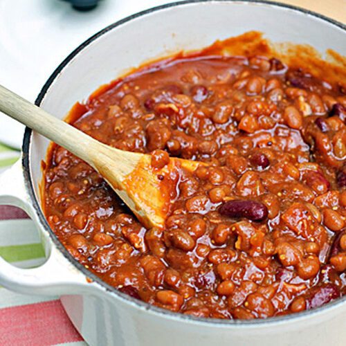 A white pot filled with thick, hearty chili made with beans and tomato sauce. A wooden spoon rests inside the pot, stirring the mixture. The pot sits on a striped cloth.