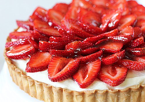 A close-up of a tart with a golden crust, topped with glossy, sliced strawberries arranged in a decorative pattern over a layer of creamy filling.