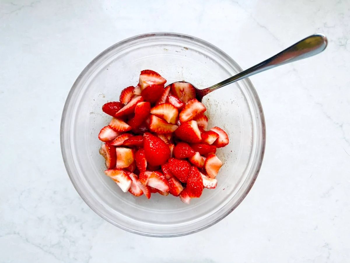 A clear glass bowl filled with sliced strawberries and a metal spoon resting inside, placed on a white marble surface—perfect for pairing with homemade Strawberry-Balsamic Ice Cream.