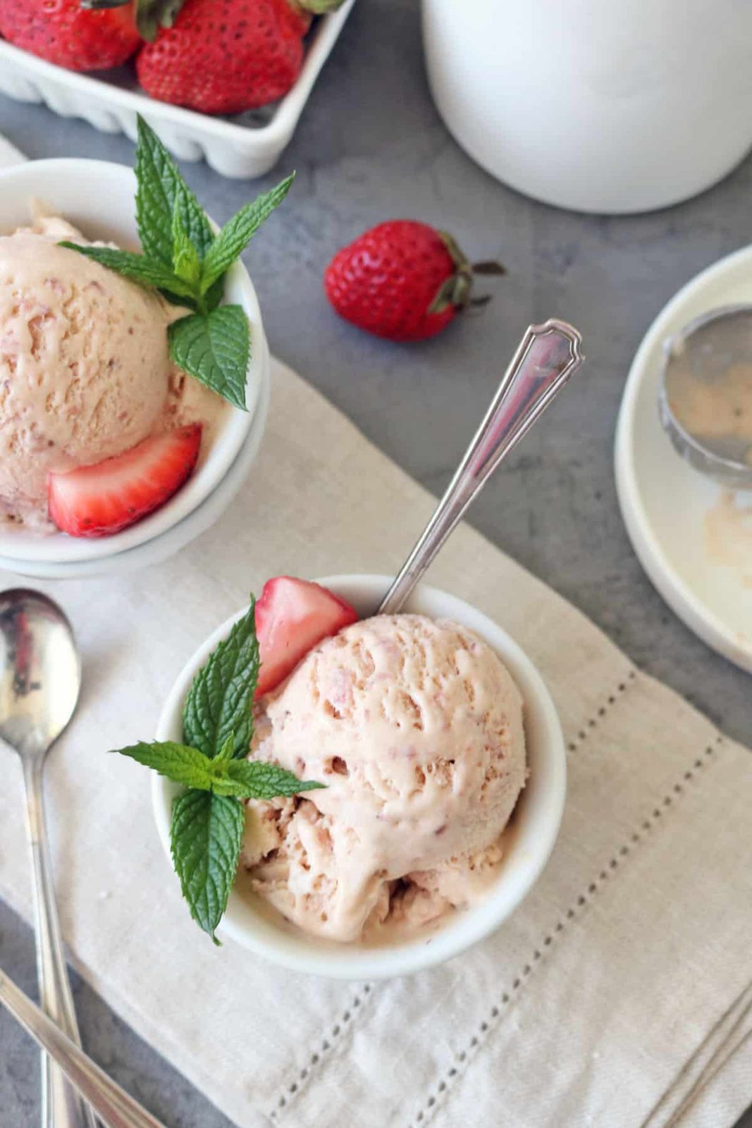 Two bowls of Strawberry-Balsamic Ice Cream garnished with fresh mint leaves and strawberry slices rest on a beige cloth, with spoons, whole strawberries, and an ice cream scoop nearby.