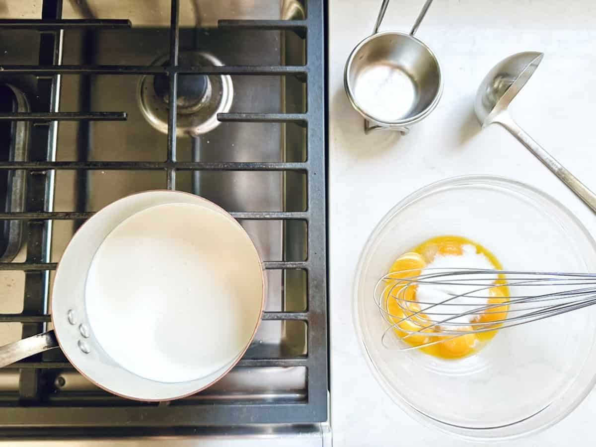 Overhead view of a stovetop with a saucepan, beside a counter holding ingredients and tools for Strawberry-Balsamic Ice Cream: a clear bowl with egg yolks and sugar, a whisk, ladle, and two measuring cups.