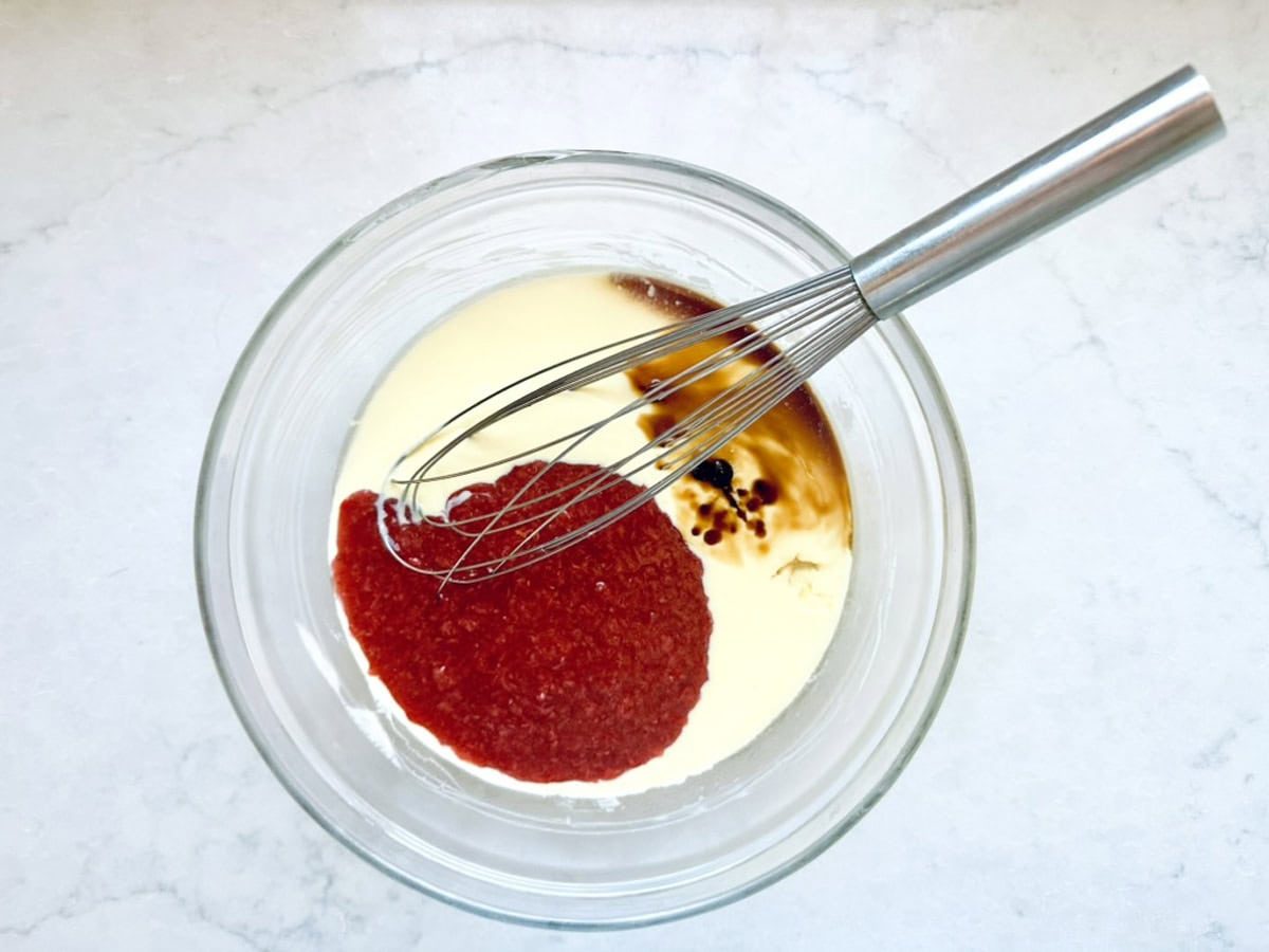 A metal whisk rests in a glass bowl containing heavy cream, bright red tomato sauce, Worcestershire sauce, and black pepper—ingredients mingling atop a white marble surface reminiscent of crafting Strawberry-Balsamic Ice Cream.