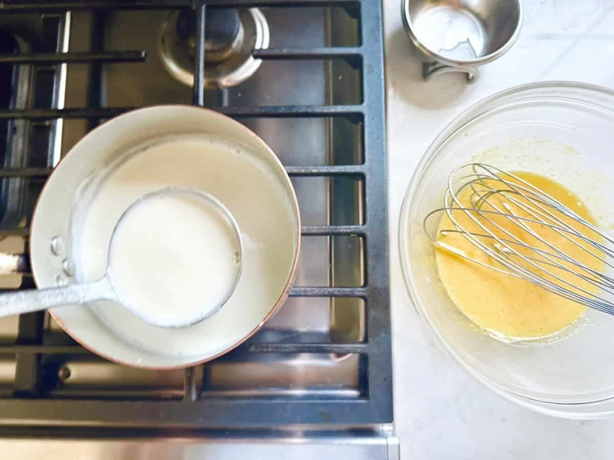 A saucepan of heated milk on a stove with a ladle inside, next to a bowl containing whisked eggs and a metal whisk—perfect prep for making creamy Strawberry-Balsamic Ice Cream on the kitchen counter.