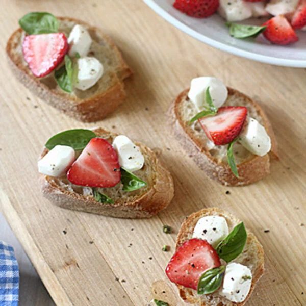 Four slices of baguette topped with fresh basil, mozzarella, and strawberry slices are arranged on a wooden board. More of the ingredients are visible in a bowl in the background. Black pepper is sprinkled on top.
