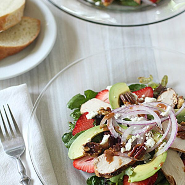 A glass plate with a salad of grilled chicken, avocado, strawberries, pecans, feta cheese, red onions, and greens. A fork rests on a napkin nearby, and a small plate of sliced bread is in the background.