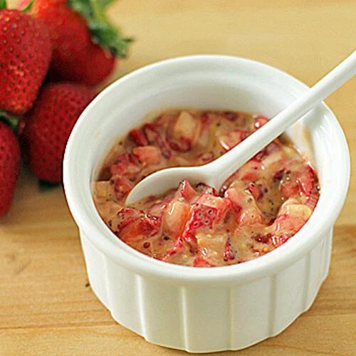 A white ramekin filled with a creamy strawberry mixture, accompanied by a white spoon. Fresh whole strawberries are placed beside the ramekin on a wooden surface.