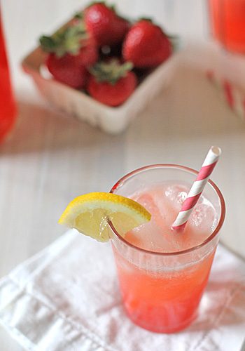 A glass of pink strawberry lemonade with ice, a striped straw, and a lemon wedge garnish sits on a napkin. In the background, a small basket of fresh strawberries is slightly out of focus.