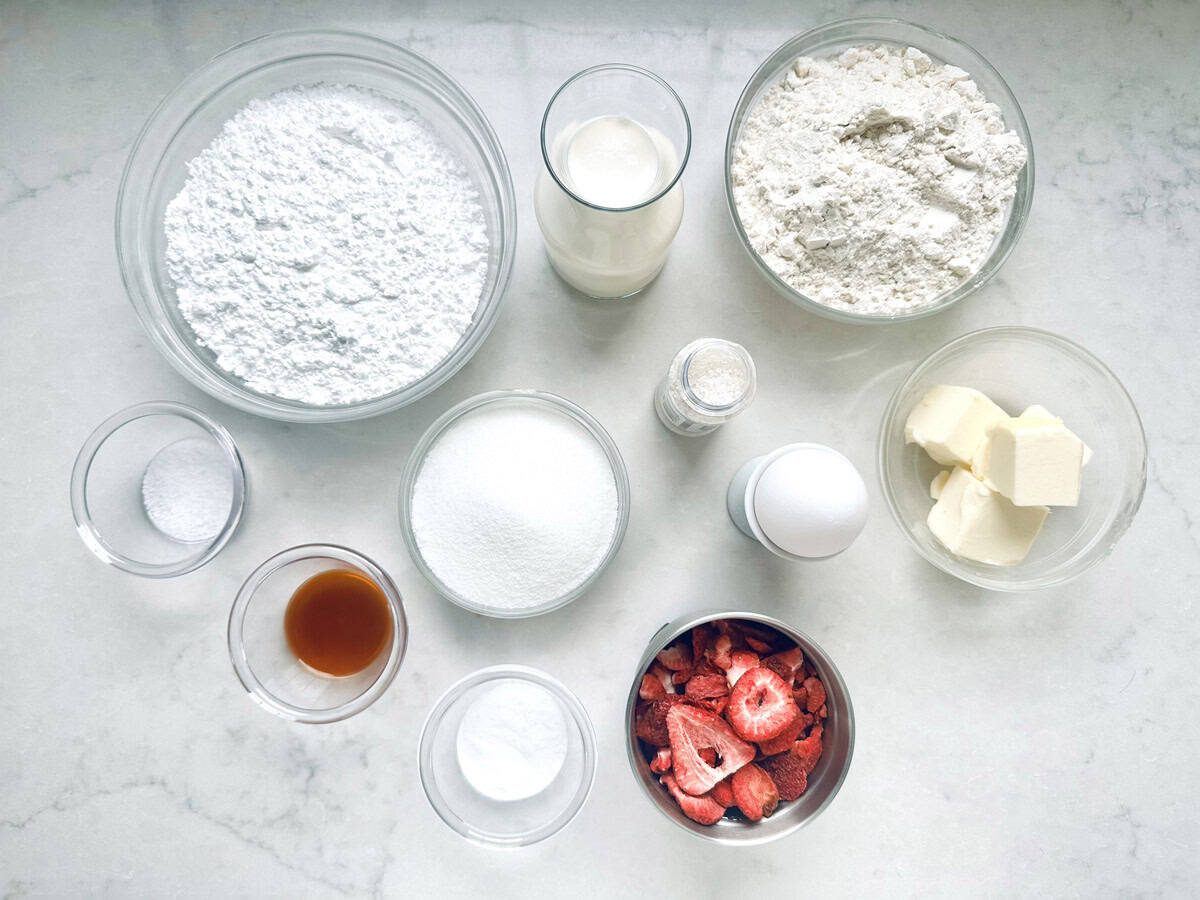 A top-down view of baking ingredients for Strawberry Shortcake Cookies in bowls and glasses, including flour, powdered sugar, milk, butter, an egg, strawberries, vanilla, salt, baking powder, baking soda, and granulated sugar on a white surface.