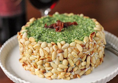 Round cheese spread coated with pine nuts on the sides, topped with a layer of green pesto and a garnish of sun-dried tomato, served on a white plate.