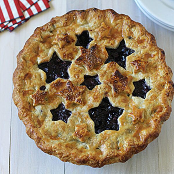 A golden-brown pie with star-shaped cutouts on the top crust, showing dark berry filling. The pie sits on a light wooden surface next to red and white napkins and stacked white plates with silver forks.