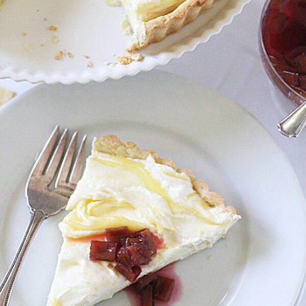 A slice of creamy lemon tart topped with rhubarb compote is served on a white plate with a fork. The rest of the tart and a bowl of compote are visible nearby.