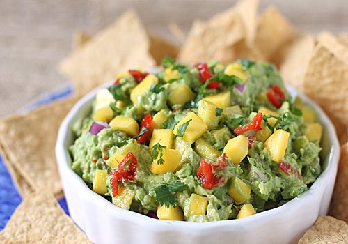 A white bowl filled with guacamole topped with diced mango, red onion, tomato, and cilantro, surrounded by tortilla chips.