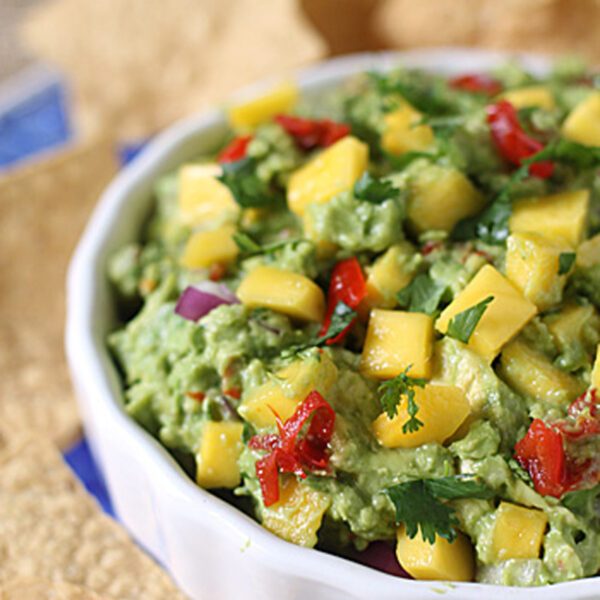 A white bowl filled with guacamole topped with diced mango, red onion, tomato, and cilantro, surrounded by tortilla chips.