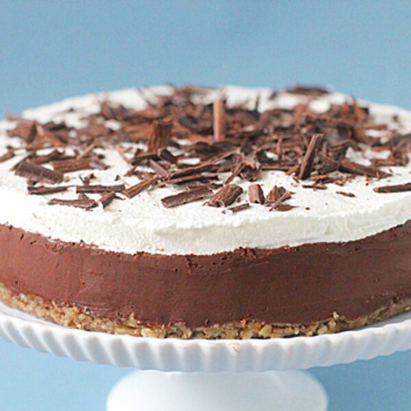 A layered chocolate and cream cake sits on a white cake stand. The cake has a nutty base, a thick chocolate layer, a fluffy white cream topping, and is garnished with chocolate shavings. The background is light blue.