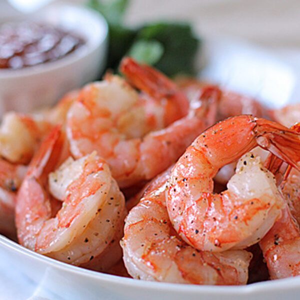 A plate of cooked, seasoned shrimp is served with a small bowl of cocktail sauce in the background. The shrimp are pink, plump, and garnished with ground pepper.