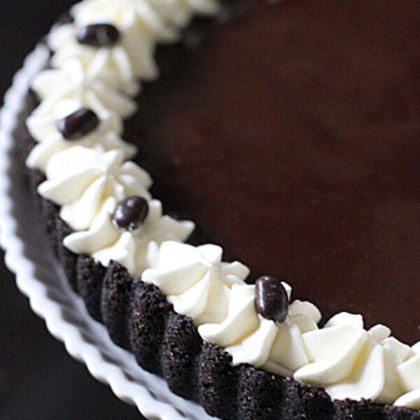 A chocolate tart on a white plate, topped with a ring of piped whipped cream and decorated with chocolate-covered coffee beans around the edge.
