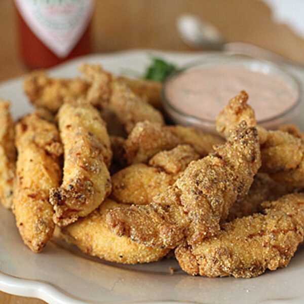 A plate of crispy, golden-brown fried catfish strips is served with a small bowl of creamy dipping sauce on the side, all set on a light-colored plate.