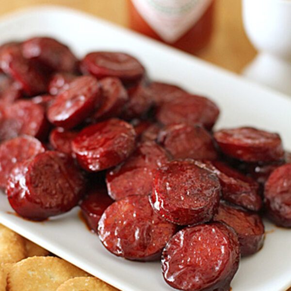A white plate filled with glazed sausage slices is shown next to a row of round crackers. A bottle of hot sauce and a small container with toothpicks are blurred in the background.