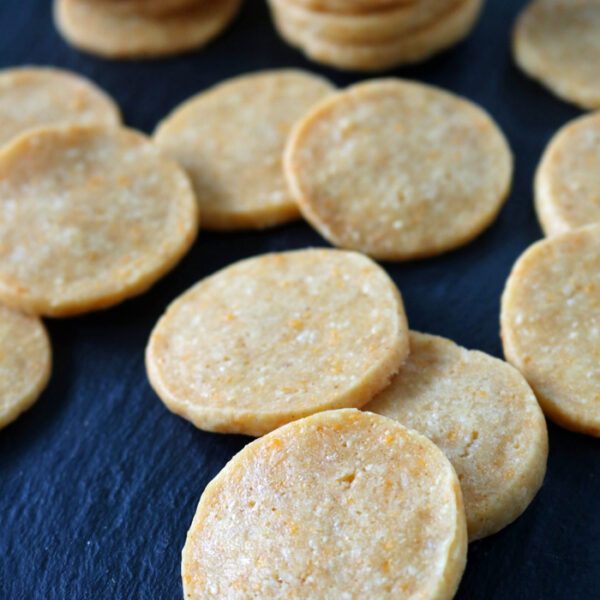 Round, golden-brown crackers are scattered and stacked in small groups on a dark, slate surface. The crackers appear crispy with a slightly textured surface.