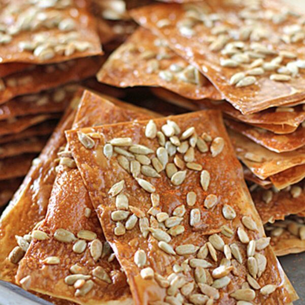 A plate stacked with rectangular, golden-brown, crispy crackers topped with sunflower seeds. The crackers appear thin and glossy, with seeds sprinkled generously on top.