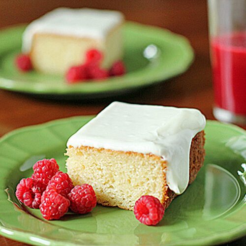 A square piece of frosted cake on a green plate with fresh raspberries, and a glass of red juice in the background. Another plate with cake and raspberries is visible further back.