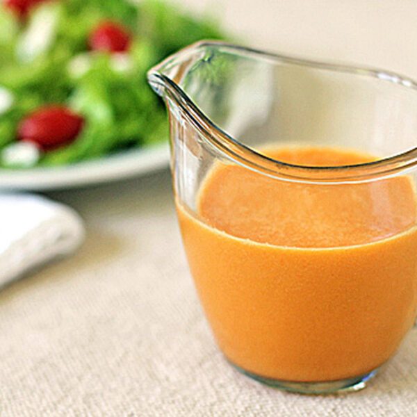 A clear glass pitcher filled with orange salad dressing sits on a beige surface, with a plate of green salad and cherry tomatoes blurred in the background.