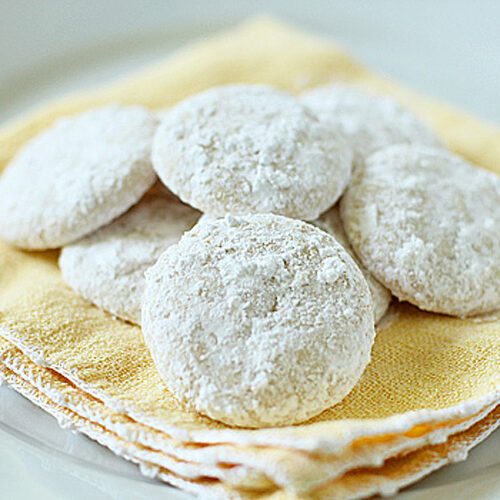 A group of round, powdered sugar-coated cookies arranged on a stack of yellow cloth napkins on a white plate.