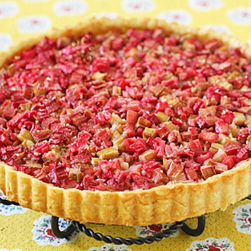 A baked rhubarb tart with a golden crust, topped with chopped pink and green rhubarb pieces, sits on a metal stand over a yellow floral tablecloth.