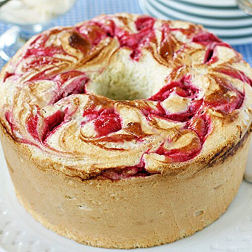 A round marble cake with swirls of raspberry on top sits on a white plate. A serving utensil is nearby, with plates and fresh raspberries in the background on a blue checkered tablecloth.