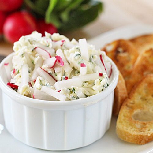 A white ramekin filled with a creamy dip garnished with chopped radishes and herbs, served on a plate with toasted baguette slices. Fresh radishes and greens are blurred in the background.