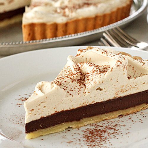 A slice of chocolate cream pie with a thick layer of whipped topping and cocoa powder sprinkled on top, served on a white plate. The whole pie is visible in the background.
