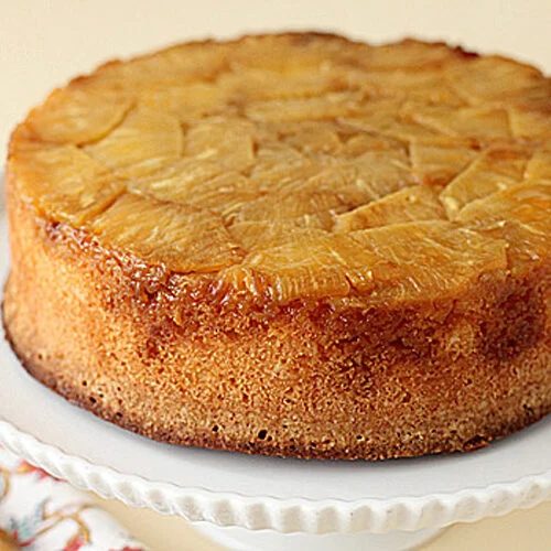 A round pineapple upside-down cake sits on a white cake stand, showing caramelized pineapple slices on top and a golden brown edge. A patterned napkin and fork are partially visible beside the stand.