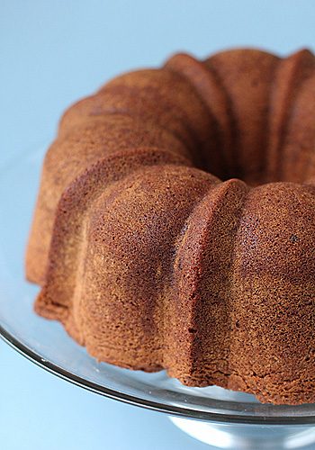 A golden-brown bundt cake with a textured surface sits on a clear glass cake stand against a light blue background.