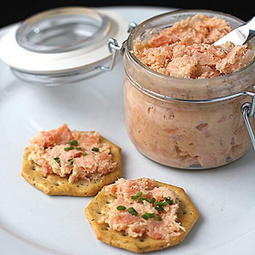 A glass jar of salmon spread with a small knife sits on a white plate next to two crackers topped with the spread and garnished with chopped chives.