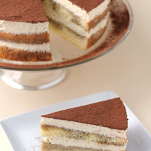 A slice of tiramisu cake with creamy layers and cocoa powder on top sits on a white plate with a fork, while the remaining cake is displayed on a glass stand in the background.