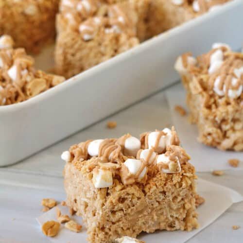 A close-up of Fluffer-nutter Treats—peanut butter Rice Krispies treats topped with mini marshmallows and a drizzle of peanut butter, with more treats in a white dish behind.