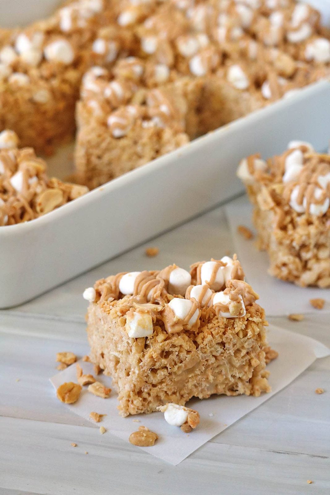 A close-up of Fluffer-nutter Treats—peanut butter Rice Krispies treats topped with mini marshmallows and a drizzle of peanut butter, with more treats in a white dish behind.