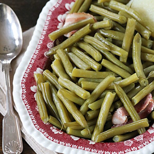 A vintage red patterned plate filled with cooked green beans and pieces of ham, placed on a white plate with a beige napkin and a silver spoon beside it.