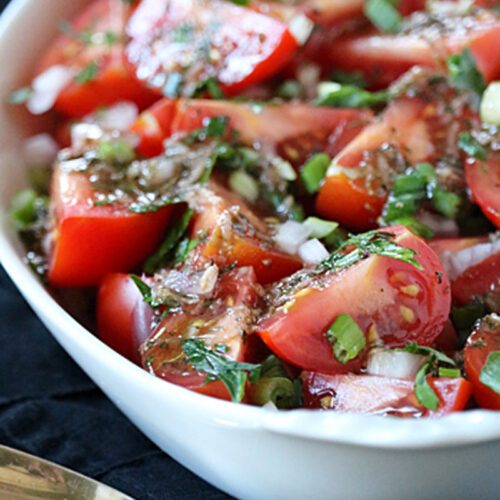 A white bowl filled with a fresh tomato salad, featuring chopped tomatoes, diced onions, green herbs, and a vinaigrette dressing, placed on a dark cloth next to a silver serving spoon.