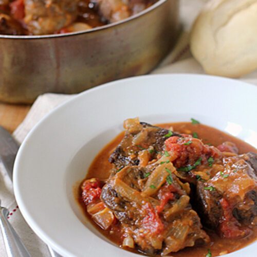 A white bowl filled with braised oxtails in a rich tomato-based sauce, garnished with herbs, sits on a napkin beside a fork and knife. In the background, a pot of oxtails and a loaf of bread are visible.